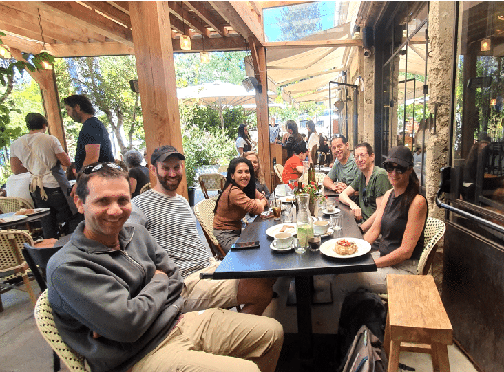 A group of people sitting at an outdoor restaurant table, enjoying food and drinks under a shaded patio.