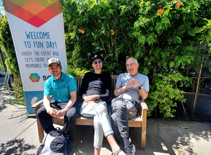 Three men sitting on a bench beside a "Welcome to Fun Day" sign in a garden setting.