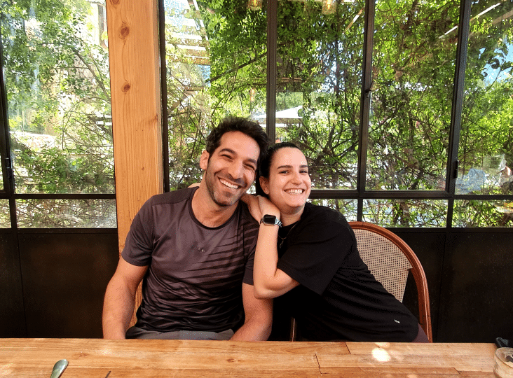 Two people sitting together at a wooden table in front of a window with greenery. They are smiling, and one of them has their arm around the other’s shoulder.