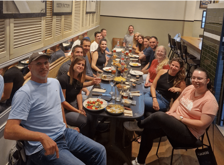A group of people seated around a long table in a restaurant, smiling at the camera. Plates of food and drinks are on the table.