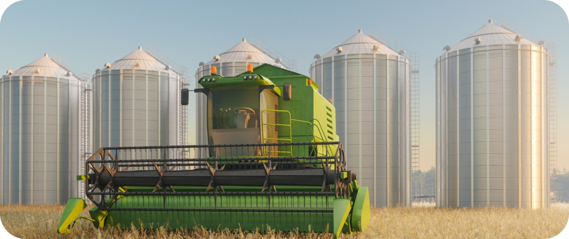 A green combine harvester in a field with five large silver grain silos in the background under a clear sky.