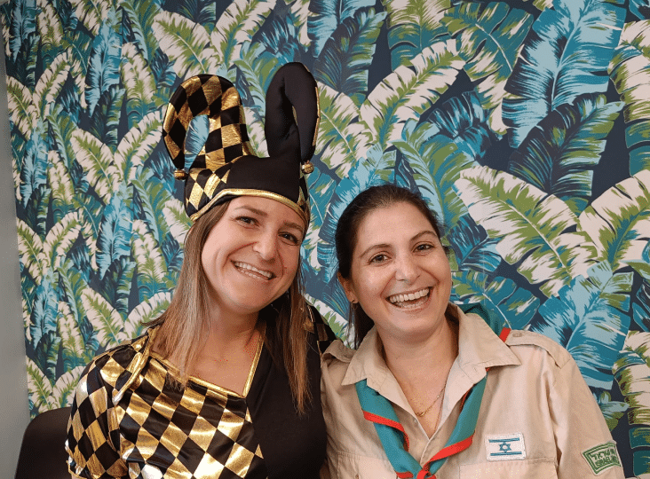Two smiling people in colorful outfits stand in front of a tropical leaf-patterned backdrop.