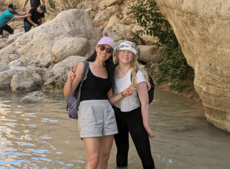 Two people stand in a shallow rocky river, smiling and flashing peace signs. Another person is visible climbing rocks in the background. They are dressed casually and wearing hats.