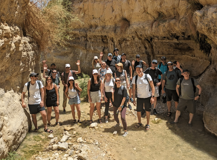 A group of people poses and waves in a rocky canyon, some standing in shallow water. They are dressed for hiking with backpacks and hats.