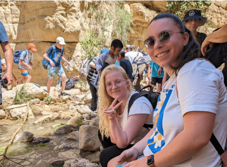 A group of people hike through a rocky area with small patches of vegetation. Two women in the foreground smile at the camera; one of them shows a peace sign.