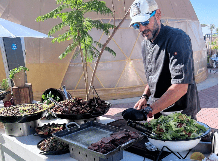 A person in a black shirt prepares and serves various dishes outdoors near a dome-shaped tent.