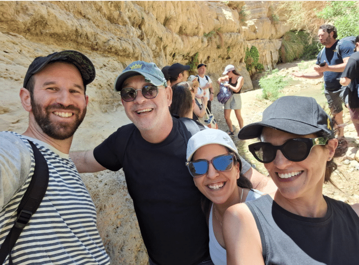 Group of people smiling and posing for a selfie outdoors in a rocky, sunlit area. They are wearing casual clothing and hats. Other people are in the background.