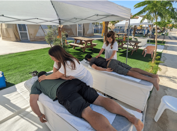 Two people receive massages on white tables under a canopy, with therapists working on their backs. The setting is outdoors, with grass, palm trees, and picnic tables in the background.