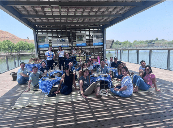A group of people sitting on a wooden deck under a pergola by the water, gathered around picnic blankets and tables.