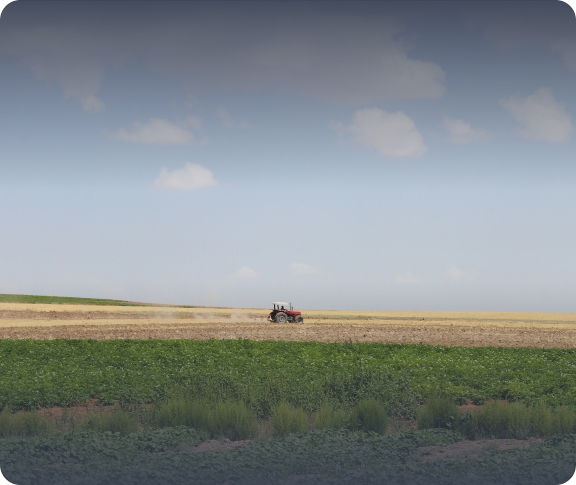 A red tractor plows a vast, open field under a partly cloudy sky.