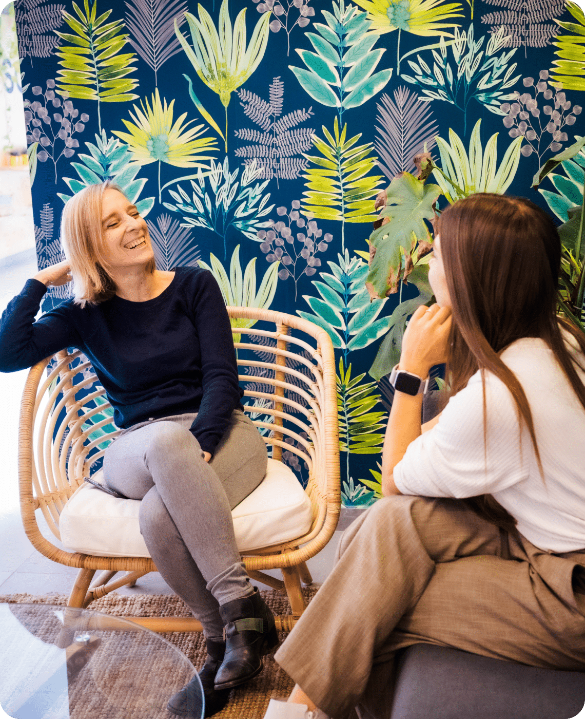Two women are smiling and chatting in a room with a vibrant plant-patterned wallpaper. One is seated in a wicker chair, while the other sits across on a cushioned seat.