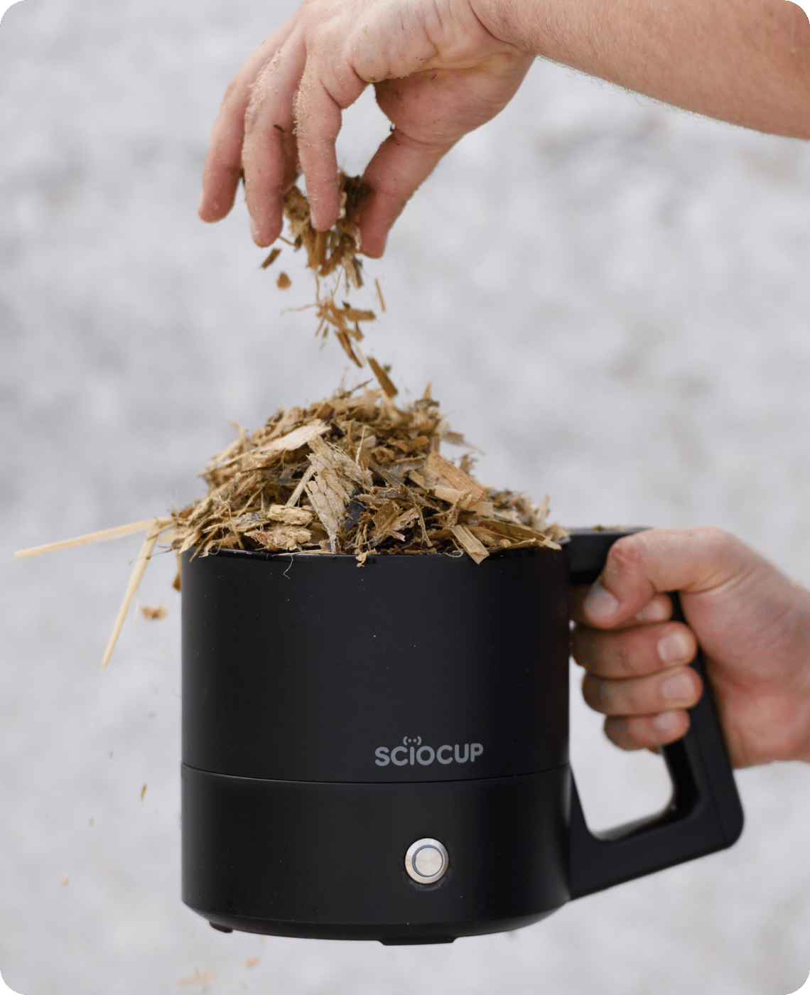 A hand is placing dried plant materials into a black ScioCup device held by another hand.