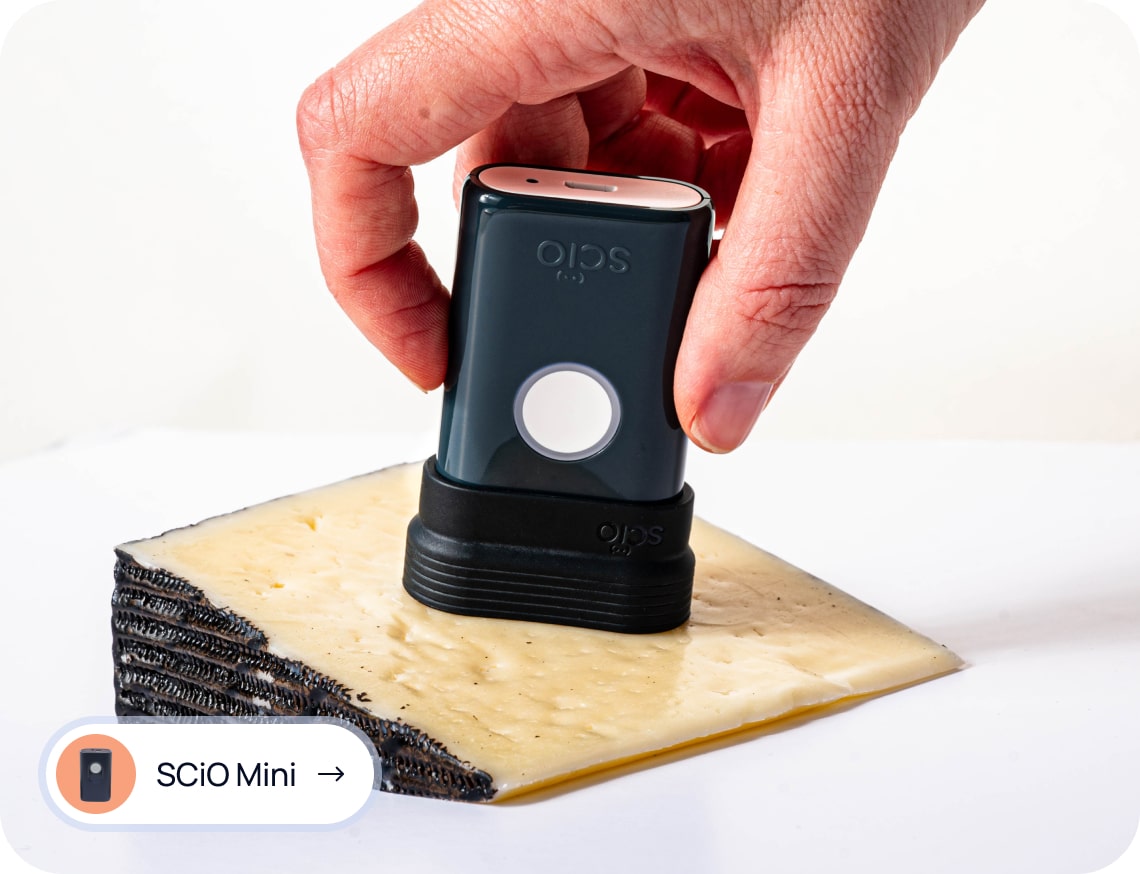 A hand holds a SCiO Mini device above a block of cheese to analyze its composition.